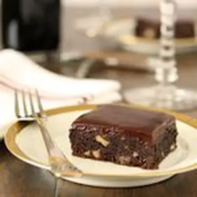 A brownie rests on a white plate with a gold rim. The brownie is filled with nuts and topped with a shiny glaze. An elegant fork rests next to it on the plate, and a napkin is hazy in the background.