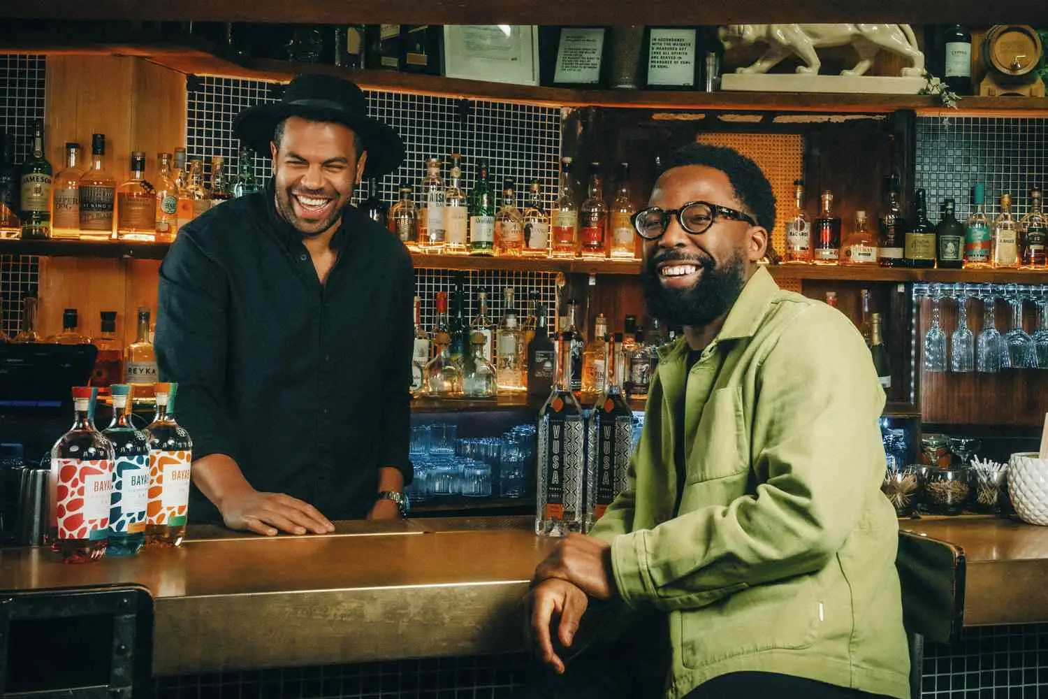 Chris Frederick and Damola Timeyin standing behind and seating in front of bar, respectively, with bottles of Bayab Gin and Vusa Vodka