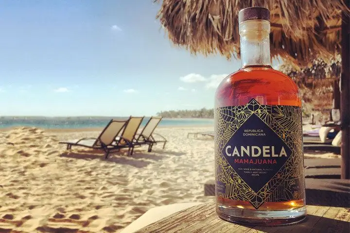 A bottle of reddish-brown Candela Mamajuana pictured on a beach beneath a straw umbrella
