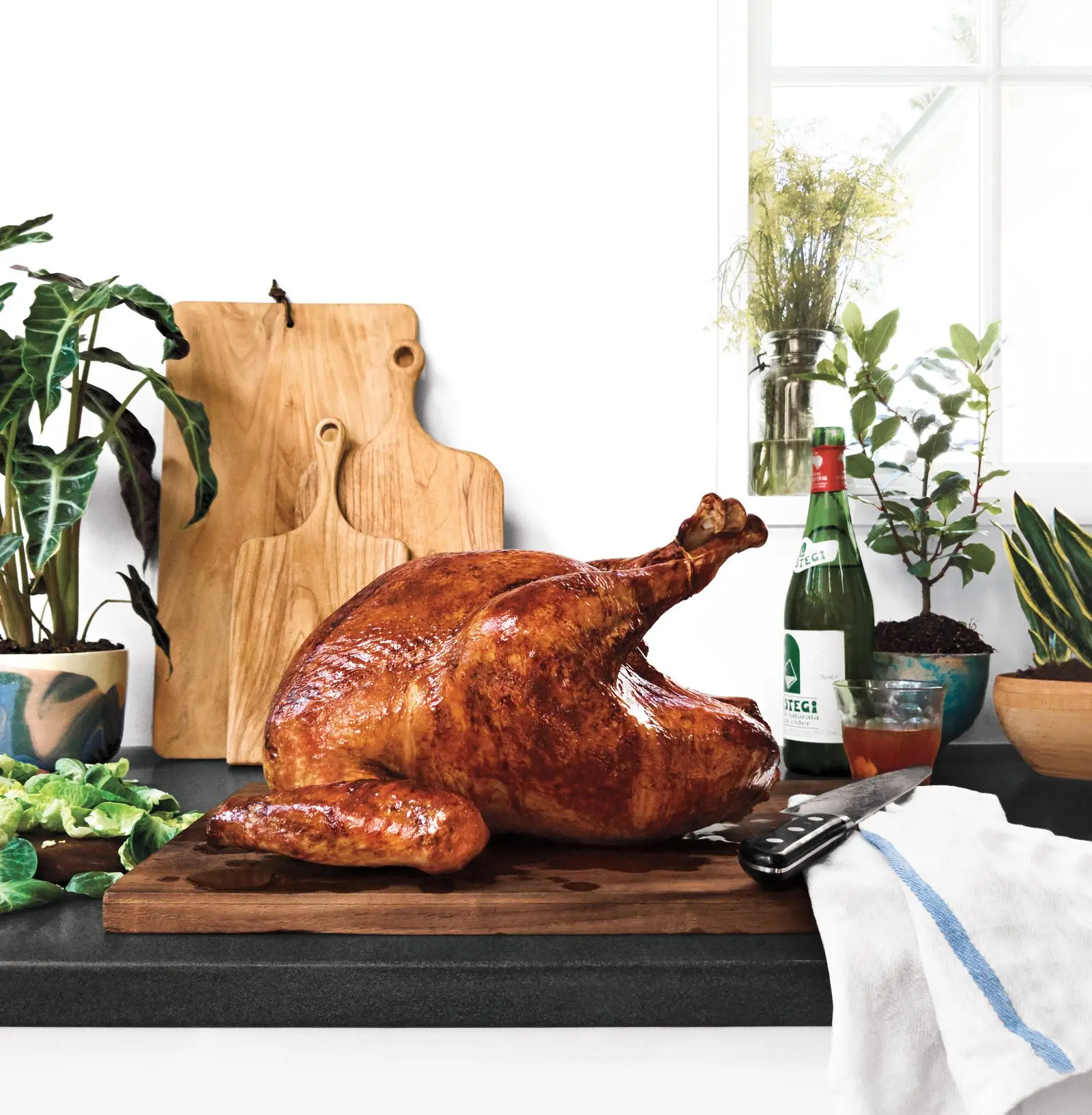 A goldenbrown roast turkey on a carving board surrounded by a bottle of wine brussels spout leaves a towel and a carving...