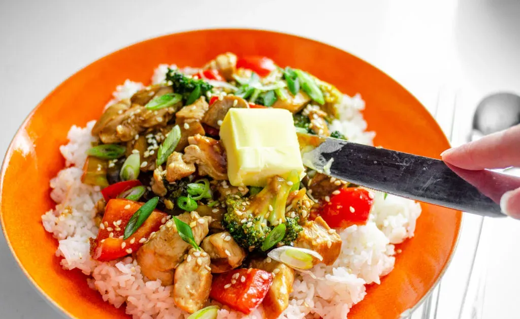 Adding a tablespoon of grass-fed butter to a bowl of healthy chicken stir fry on top of white rice.' title='Healthy Chicken Stir Fry Recipe