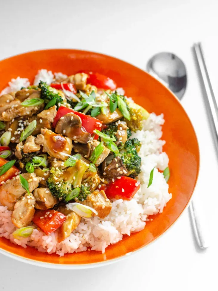 A bowl of healthy chicken stir fry on top of a bed of rice with a spoon and chopsticks next to it.' title='Healthy Chicken Stir Fry Recipe