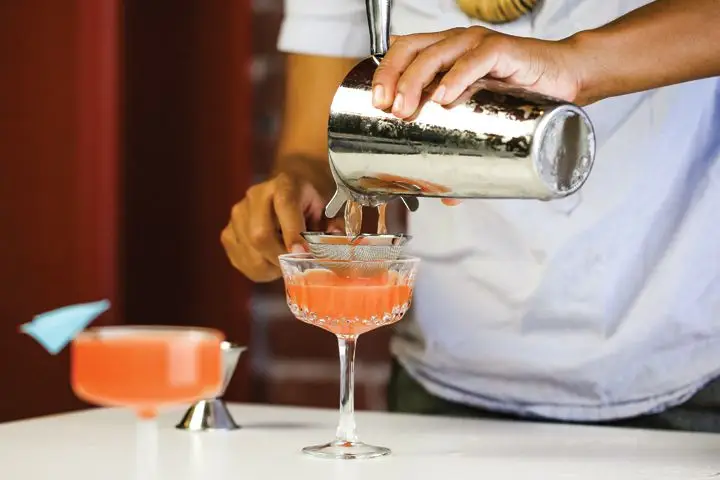 A bartender fine-straining a deep orange Paper Plane cocktail into a coupe glass