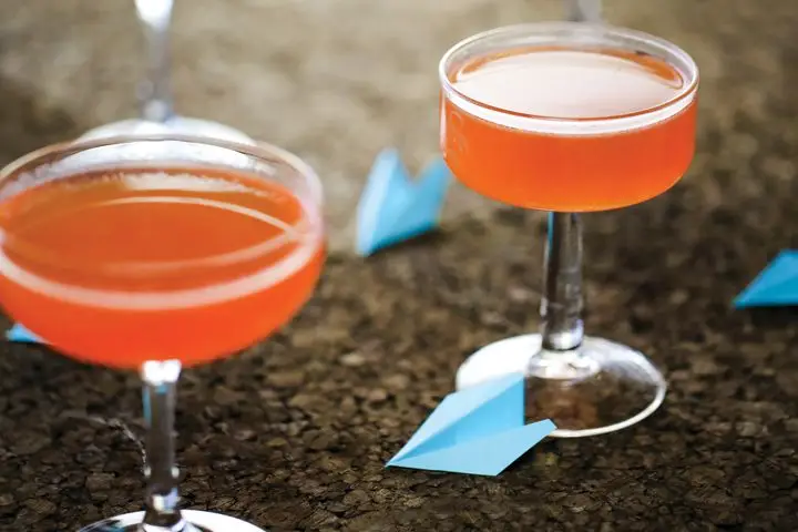 Several Paper Plane cocktails lined up with small blue paper airplane garnishes scattered around