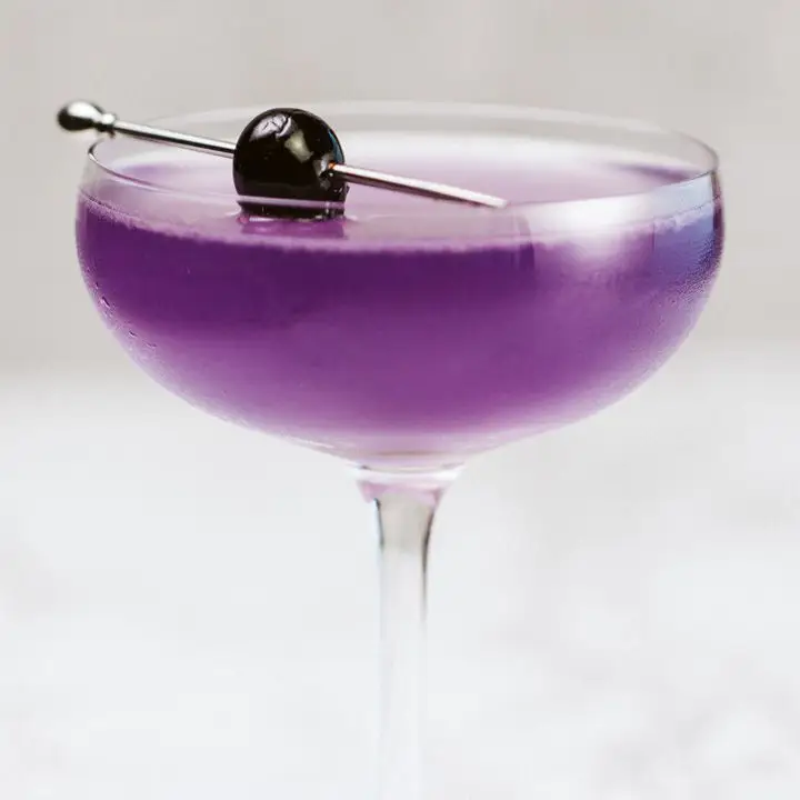 A vividly purple Aviation in a delicate coupe glass is garnished with a dark cherry, pierced on a silver pick