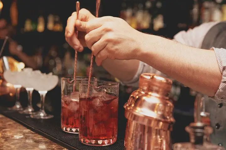 A bartender stirs two mixing glasses of Manhattans on a stone bar top with chilled coupes in the background.