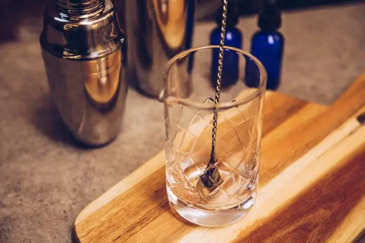 A clear stirring glass with an etched diamond pattern sits on a wooden kitchen paddle. A long-handled bar spoon rests in the glass, and next to it are some shakers and stoppered glass bottles.