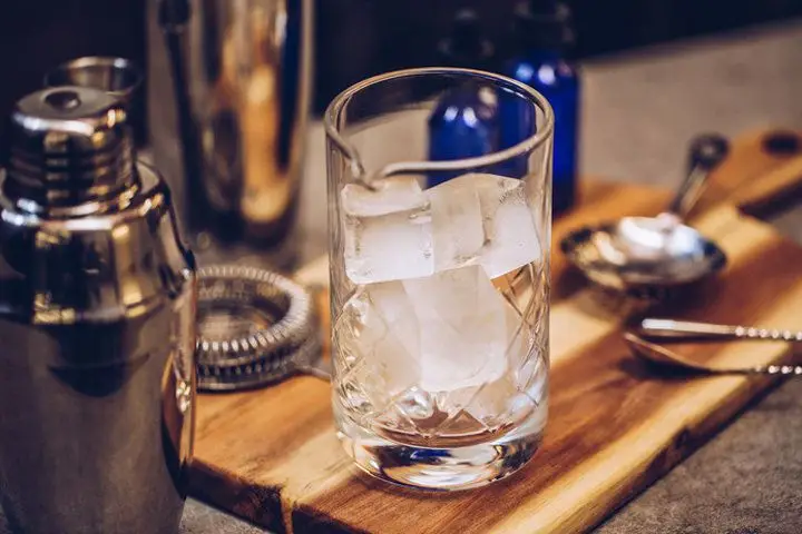 Centered in the photo is a clear mixing glass filled with large ice cubes. The mixing glass is on a wooden cutting board surrounded by other bar accoutrements including bar spoons, strainers, and shaker tins. 