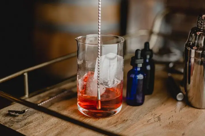 A clear mixing glass with smooth walls is filled with two large ice cubes and a bright red spirit. The bottom end of a twisted bar spoon is extended into the glass, and two small blue bottles stand next to it. All of it is placed on a wooden bar tray with a bronze railing around it. 