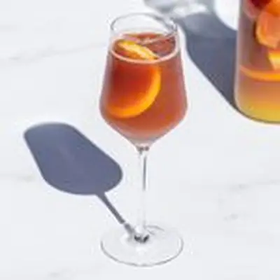 Red Sangria in wine glass with ice and orange slice on white marble, with pitcher of Sangria in background
