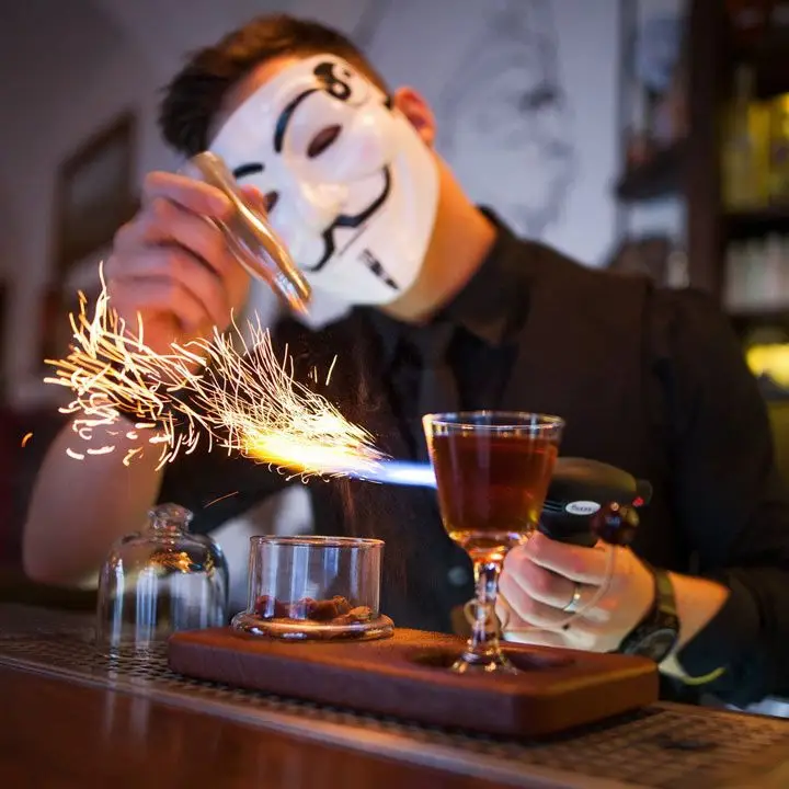 A cocktail at AnonymouS Bar in Prague. The bartender is wearing an Anonymous mask and sparks are coming from a blowtorch near a cocktail glass