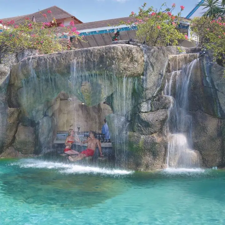 Crystal Cove swim-up bar in Barbados. Two patrons are sitting on a barstool near a bar which is tucked under a rock outcropping. A waterfall pours over the area in front of the guests