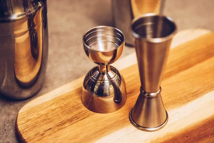 Two different jigger styles, both copper-gold in color, placed on a wood serving board