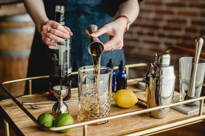 A bartender behind a wooden bar cart pours a brown spirit into a beveled mixing glass from a Japanese jigger