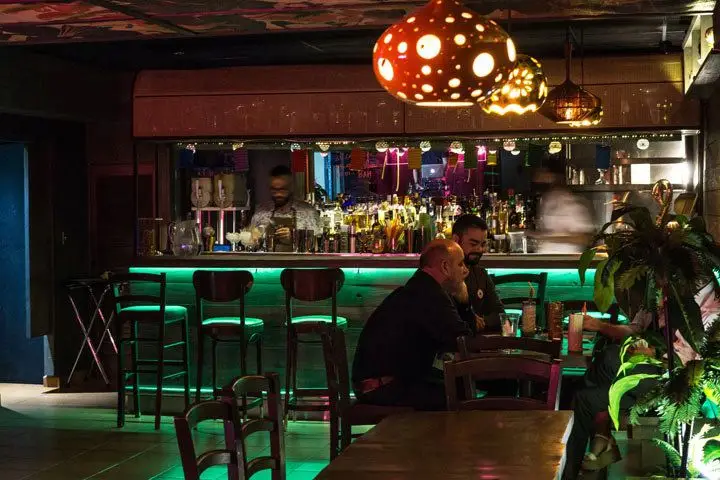 A dimly-lit tropical-meets-Tiki space with green neon illuminating the bar next to several people sitting at a table