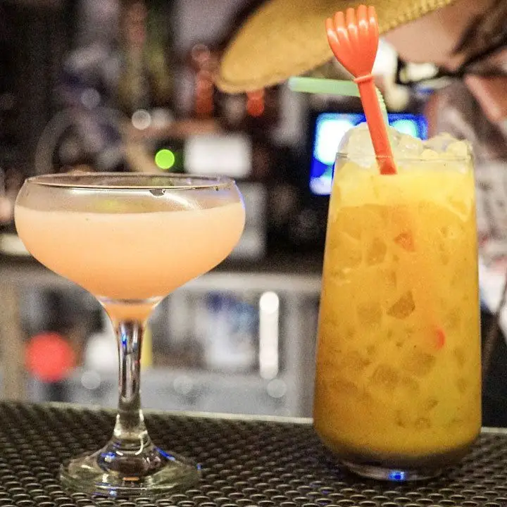 A light pink cocktail in a coupette next to a highball containing a shaken orange cocktail on crushed ice and garnished with an orange swizzle stick and a green straw