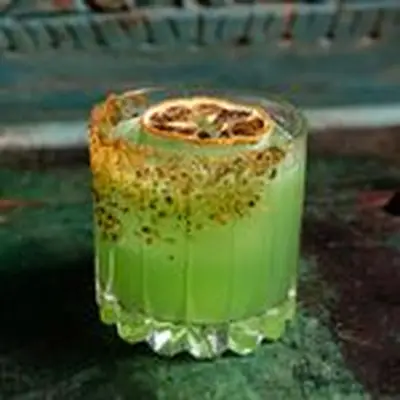 A faceted rocks glass rests on a green bar top. The glass is partially coated in chile salt, and the drink within is a vivid green. It’s garnished with a dehydrated blood orange wheel. 