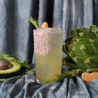 A yellow-hued cocktail in a clear Collins glass with a generous pink salt garnish on the side of the glass and a variety of fruits and vegetables surrounding the drink