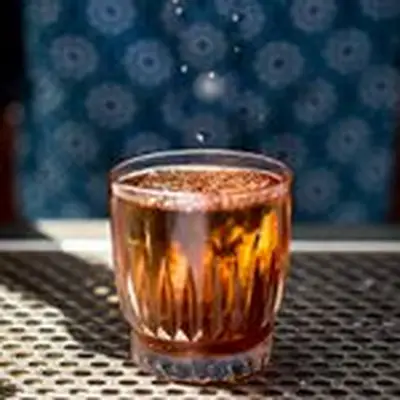 An elegant, faceted Old Fashioned glass sits on a perforated sheet of metal behind a bar. The drink within is red-gold, and a large ice cube is garnished with granulated grasshopper salt. A few flakes of the salt fall from above.