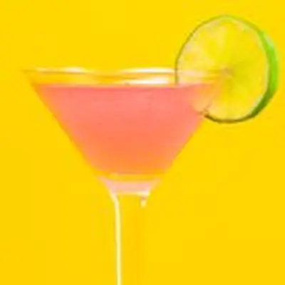 A vivid pink Cosmopolitan in a classic Martini glass is garnished with a whole lime wheel. The background is bright yellow