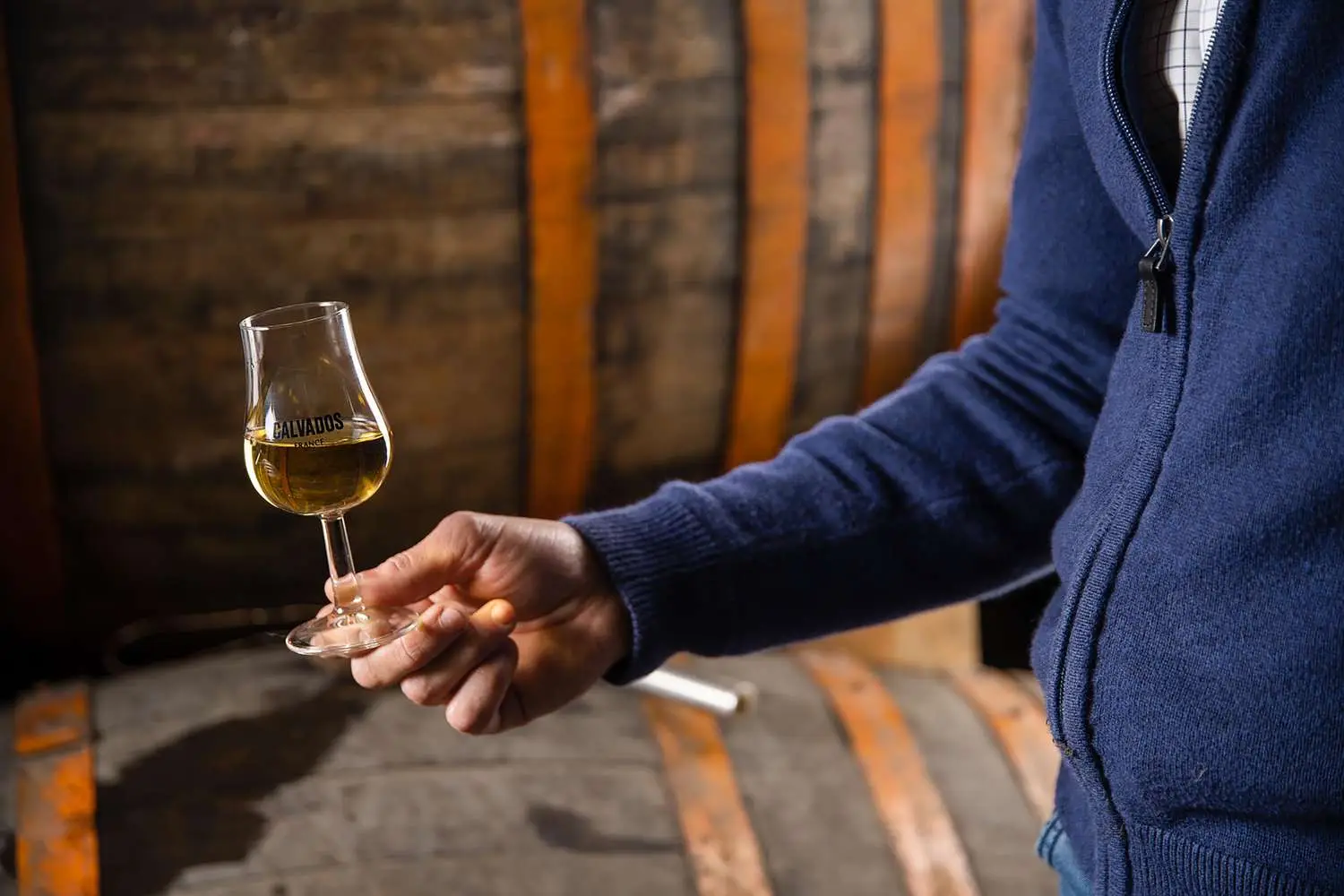 Out of frame man holding glass of Calvados with oak barrels in background