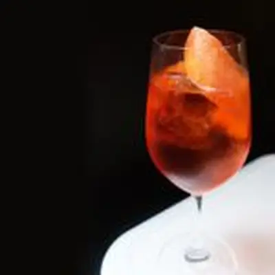 On a white table with a solid black backdrop sits a loan, high walled wine glass. It’s filled with a red-orange liquor and a few ice cubes, with a long grapefruit peel garnishing it.