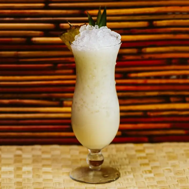 A tall and shapely glass full of crushed ice and creamy off-white Piña Colada stands on a wicker floor. Behind it, dark wood lines frame its curved shape