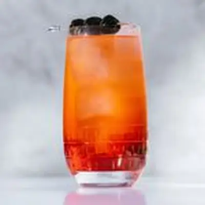 Shirley Temple in a rounded highball glass. The drink has only been lightly stirred so the red color is more intense at the base of the drink. A small metal skewer holds three Luxardo cherries and sits across the lip of the glass