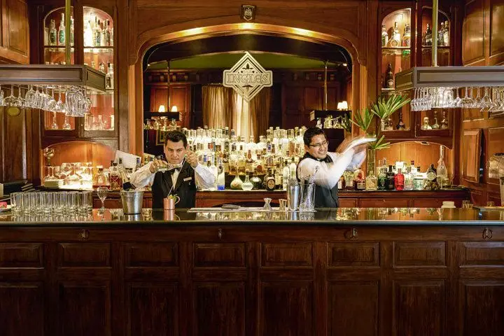 Two bartenders in action making drinks behind a bar made of deep brown paneled wood and a stone top