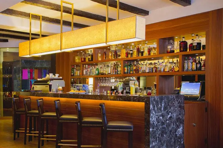 A marble-topped bar illuminated by a warm, rectangular chandelier and bottles on wood shelves