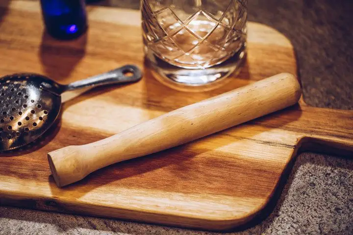 A wooden muddler placed on a wooden cutting board next to a mixing glass and strainer