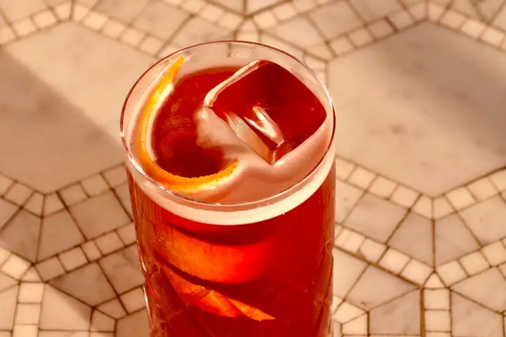 An effervescent cherry-red drink in a highball glass with a long citrus twist and a geometric tile background
