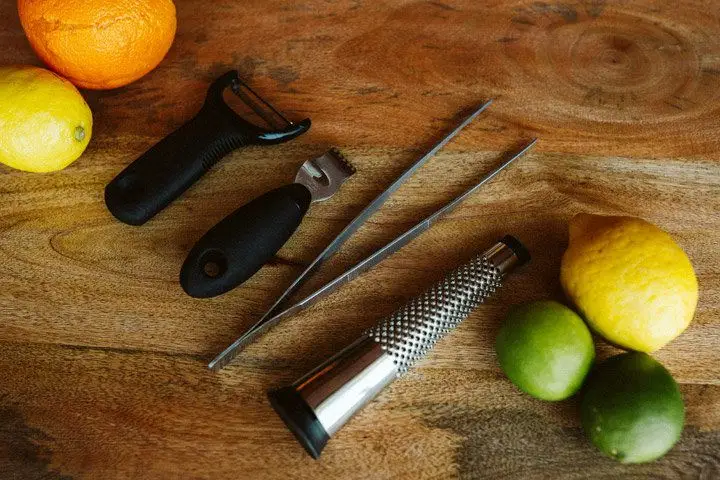 A fruit peeler, citrus planer, large tweezers, and a small citrus grater rest on a wooden tray between an array of citrus fruits.