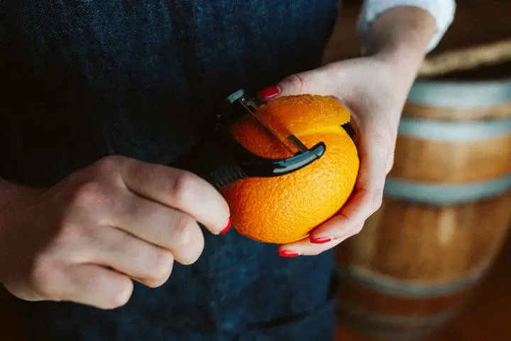 Using a wide peeler, a pair of hands with painted nails removed a large swath of peel from an orange