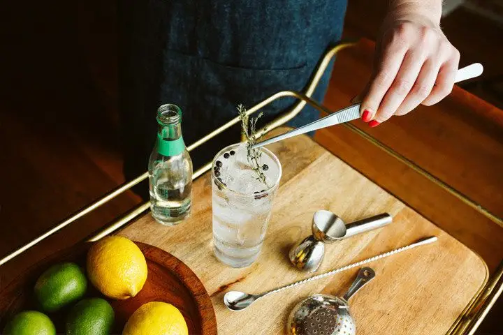 A hand with red painted nails uses large tweezers to gingerly place a sprig of thyme upon a tall glass of clear liquid and ice with juniper berries floating in it. Next to it, on a wooden tray, is an open bottle, a bowl of lemons and limes, and some other bar gear. 