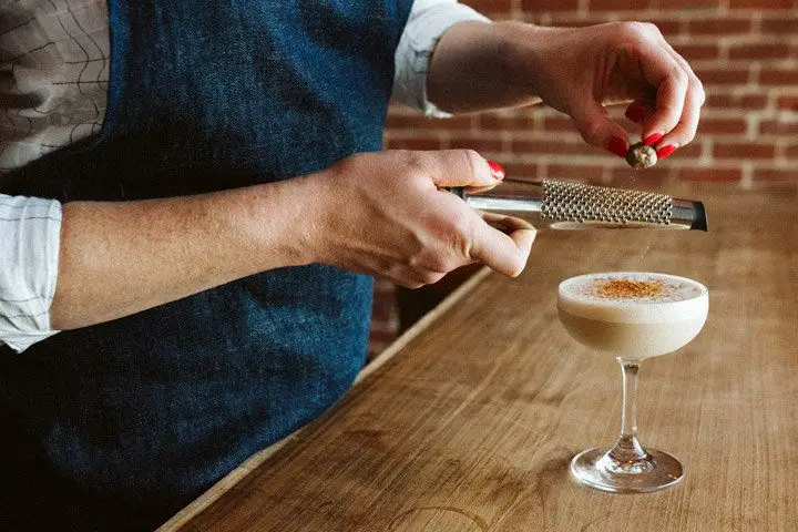 A woman wearing a navy apron is mostly out of the shot, except for her torso, arms, and hands, whose fingers are painted bright red. She grates a chunk of nutmeg over a frothy, light brown cocktail in a coupe glass. 