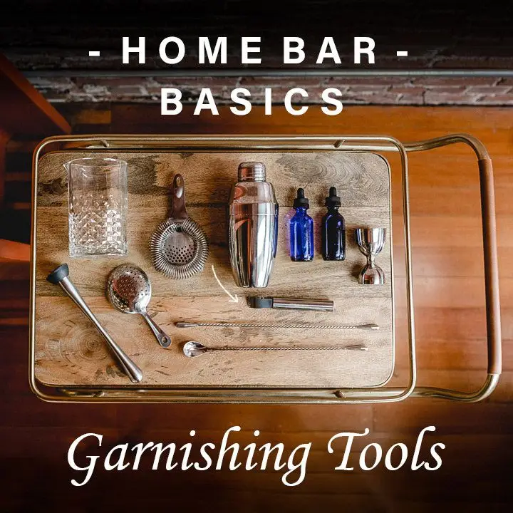 A cocktail mixing glass, two strainers, a shaker, peeler, two stoppered glass bottles, a metal jigger, two long-handled bar spoons, and a muddle rest on an elegant wooden bar tray with brass handles. A text overlay reads Home Bar Basics, Garnishing Tools.