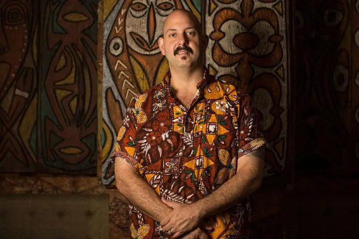 A man in a short-sleeved button down Tiki shirt standing in front of a Tiki-themed mural