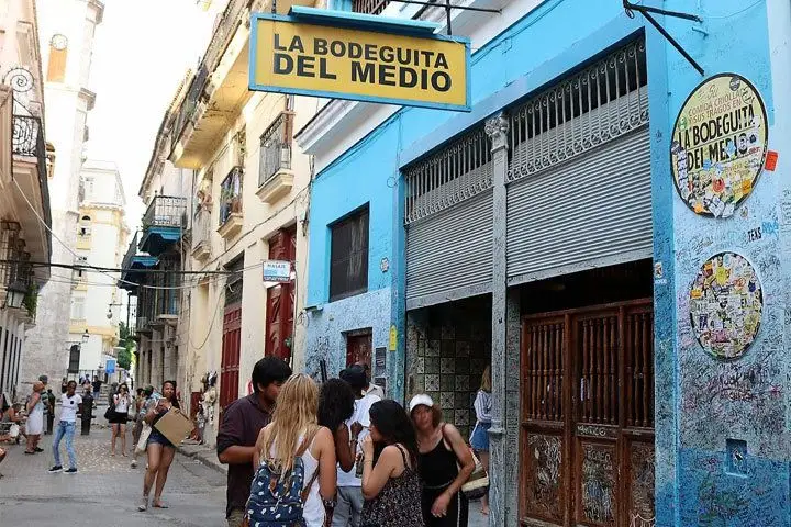 A small crowd gathers in the day time outside of La Bodeguita Del Medio, a bar in Cuba. Its walls are pale blue and covered in markings. 