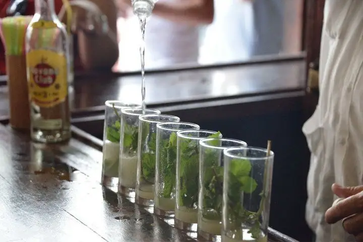 Seven glasses with sugar and mint are lined up on a sunny bar. A man, mostly off camera, pours a long stream of soda water into one of them. 