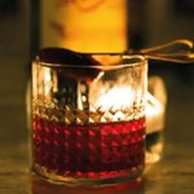 A faceted rocks glass sits on a bar top in front of a candle which illuminates the vivid red drink within. The glass is garnished with a Medjool date on a skewer.