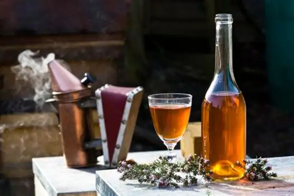 A full glass and mostly filled glass bottle filled with golden-hued wine sit on a white table, their bases surrounded by heather. In the background, out of focus, a copper devices spills smoke