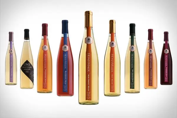 Nine narrow bottles of mead, each a different hue, are arranged in a V formation on a black white background. Each has a different colored stripe of paper running down its front, with the word Honeymaker, and the style of the mead written on it