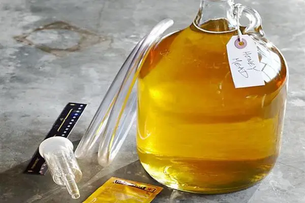 A large carboy of amber mead sits on a concrete surface. A tube leans against it, and various brewing equipment, including a bag of yeast, lay on the floor. Around the carboy