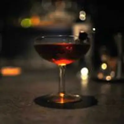 A wide, shallow coupe with etchings on the glass sits in a dark bar on a gray counter. The drink within is dark and crimson.