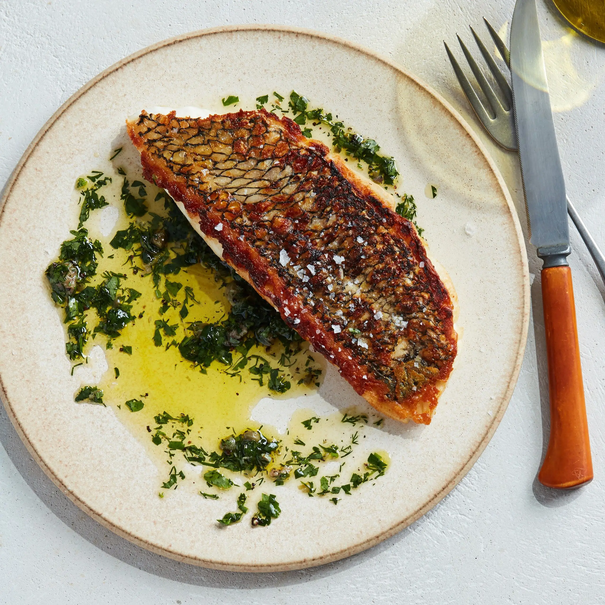 A white plate with crispyskinned fish herb sauce and a fork and knife next to the plate.