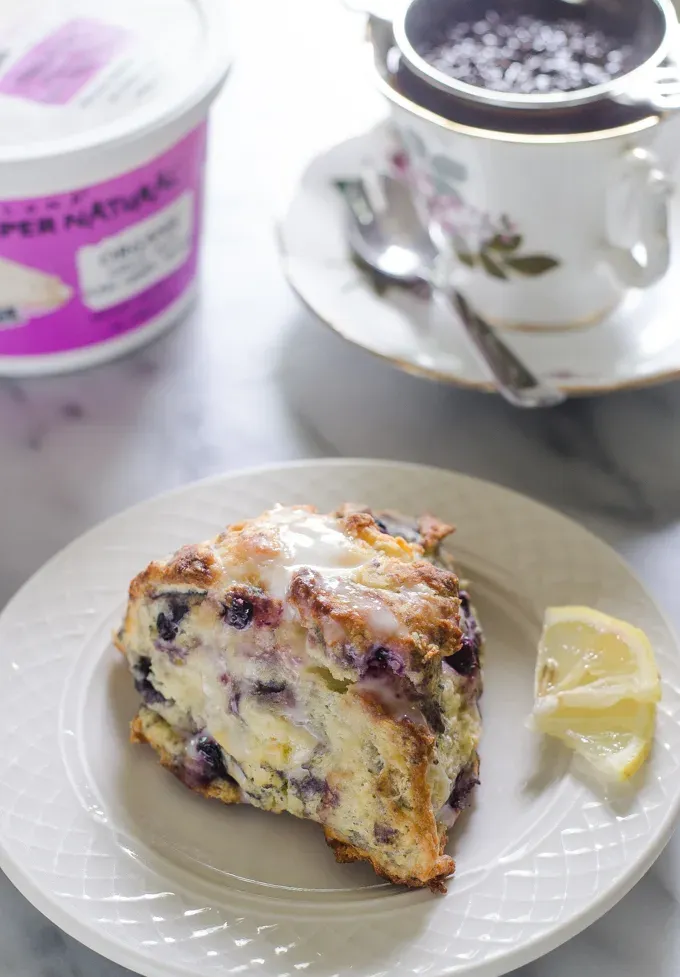 Blueberry scones on a dessert plate with small slices of lemon off to the side and a cup of tea and a tea pot in the background.' title='Easy Blueberry Greek Yogurt Scones Recipe with Lemon