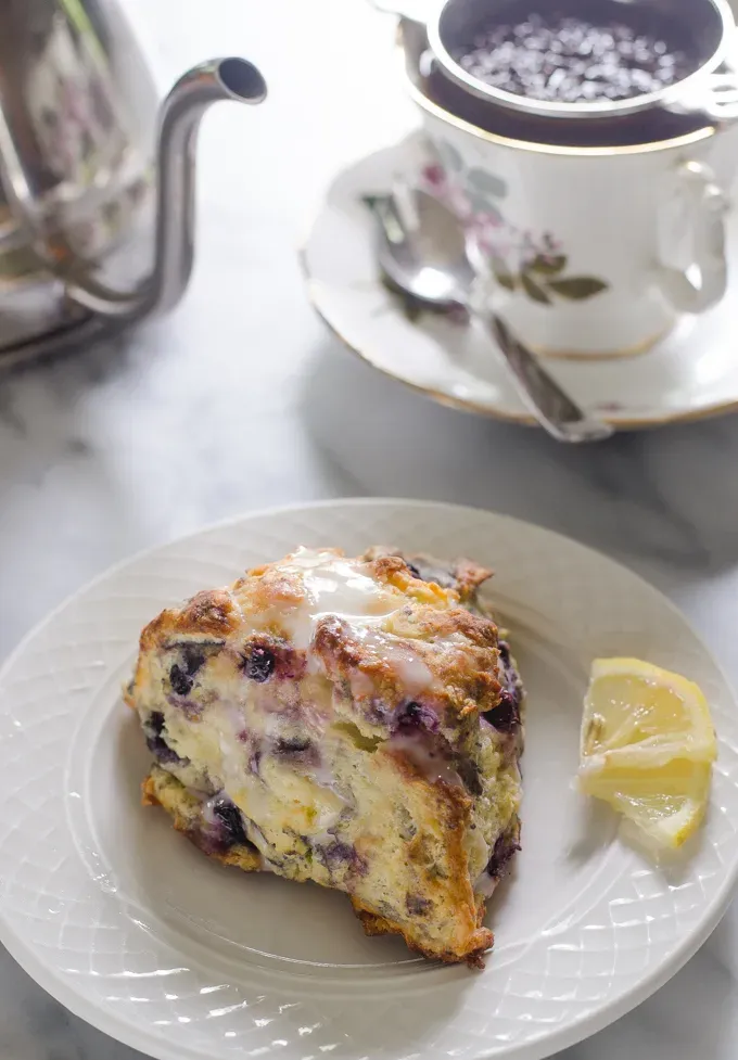 A blueberry Greek yogurt scone on a small plate with mini lemon slices next to it and a teapot and cup of tea in the background.' title='Easy Blueberry Greek Yogurt Scones Recipe with Lemon