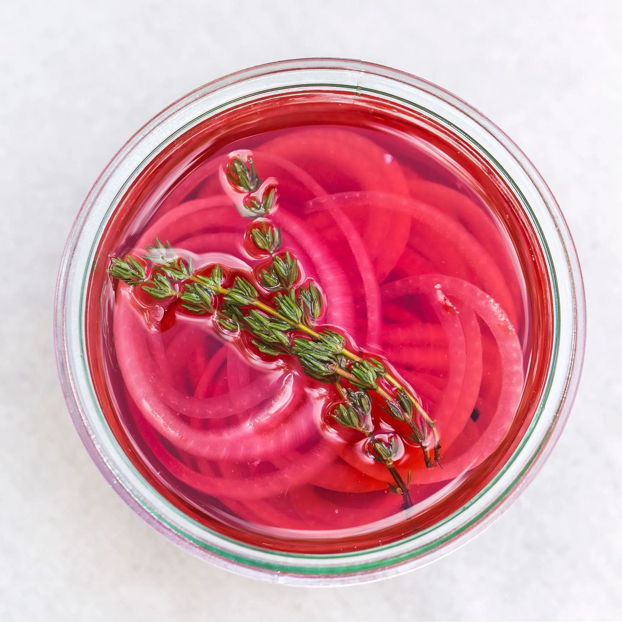 A jar of pickled red onions with thyme leaves is on a table.
