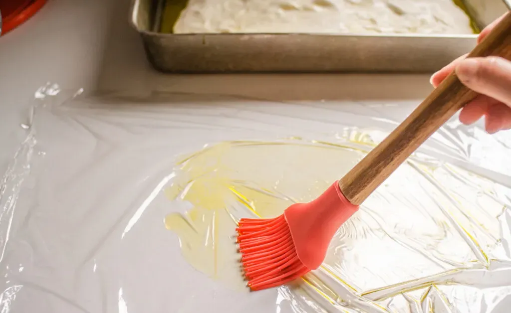 Greasing a piece of plastic wrap with olive oil to put on top of the focaccia dough.' title='Sourdough Focaccia Recipe